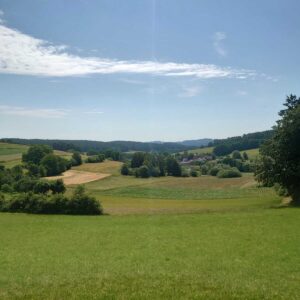 Weiter Blick in die Natur. Sonnenschein, grüne Wiesen und Hügel. Bäume und Felder sind zu sehen.
